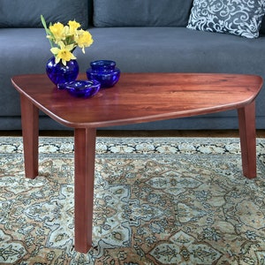 Könnte beinhalten: Ein dreieckiger Couchtisch aus dunklem Holz mit abgerundeten Kanten steht auf einem gemusterten Teppich vor einem blauen Sofa. Der Tisch hat drei Beine und ist mit einer Vase mit gelben Blumen und blauen Glasschalen dekoriert.