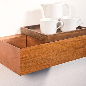 Può includere: Una mensola galleggiante in legno con un cassetto integrato. Un vassoio di legno più piccolo sopra contiene due tazze bianche e una teiera bianca. La mensola è realizzata in legno dai toni caldi ed è montata su una parete bianca.