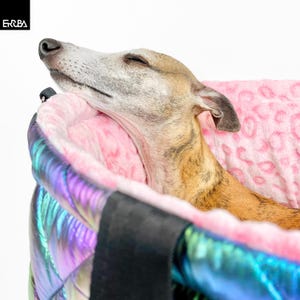 Op de afbeelding: Een hond rust in een gewatteerde dierenvervoerder. De drager heeft een roze luipaardprint interieur en een glanzende, iriserende buitenkant. De hond is lichtbruin en wit, met gesloten ogen.