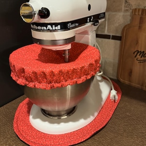May include: A white KitchenAid Classic Plus stand mixer with a stainless steel bowl. The bowl is covered by a red fabric cover with a small floral pattern, and the mixer sits on a matching red fabric mat with the same floral print design.