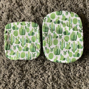 May include: Two white fabric chair cushions with a green cactus pattern. The cushions are on a brown shag carpet.