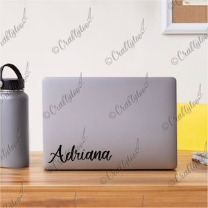 Op de afbeelding: Een zilveren laptop met de naam "Adriana" in zwarte, cursieve letters op de deksel. De laptop staat op een houten tafel.