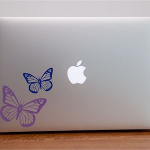 Puede incluir: Un portátil plateado con dos pegatinas de mariposas. Una mariposa es azul y la otra es morada.
