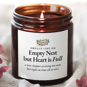 May include: A lit candle in a brown glass jar with a white label featuring a bird nest illustration and the text: Smells like an Empty Nest but Heart is Full, a new chapter arriving too soon but right on time all at once.