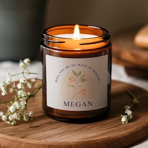 Peut inclure: Une bougie en verre brun avec une étiquette blanche qui dit "Veux-tu être ma demoiselle d'honneur ?" avec un motif floral et le nom "Megan".