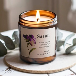 May include: A brown glass candle jar with a white label. The label has a purple flower illustration and the text "Sarah Wishing you lots of love and joy on your birthday Violet FEBRUARY".