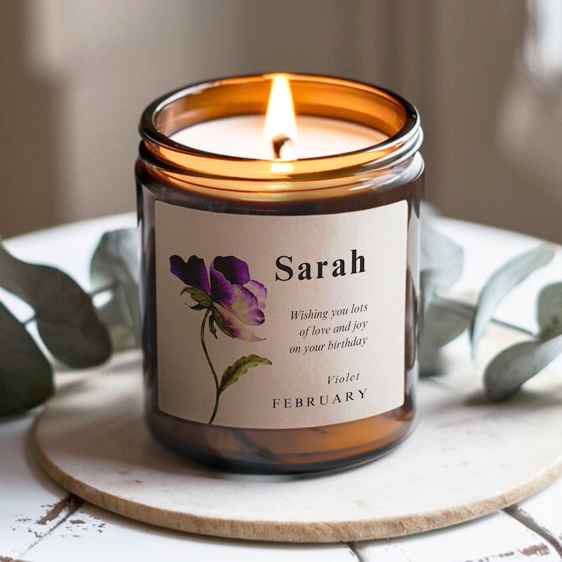 May include: A brown glass candle jar with a white label. The label has a purple flower illustration and the text "Sarah Wishing you lots of love and joy on your birthday Violet FEBRUARY".