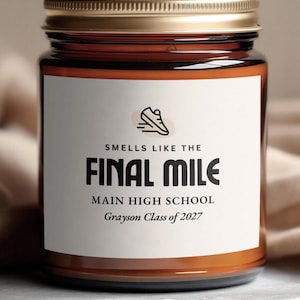 May include: A brown glass candle jar with a gold metal lid and a white rectangular label. The label features a black running shoe icon and the text: SMELLS LIKE THE FINAL MILE, MAIN HIGH SCHOOL, Grayson Class of 2027 in black lettering.