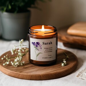 May include: A brown glass candle with a white label. The label has a purple flower illustration and the text "Sarah Wishing you lots of love and joy on your birthday Violet FEBRUARY".