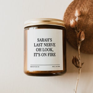 Op de afbeelding: Een bruine glazen kaarsenpot met een gouden deksel en een wit etiket. Het etiket luidt "SARAH'S LAST NERVE OH LOOK, IT'S ON FIRE." De kaars is 227g en heeft een brandtijd van 40 uur.