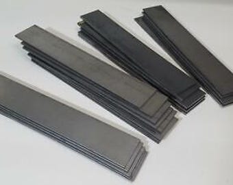 May include: A stack of five flat, rectangular metal bars, each about 12 inches long and 1 inch wide. The bars are a dark gray color and have a slightly rough texture.