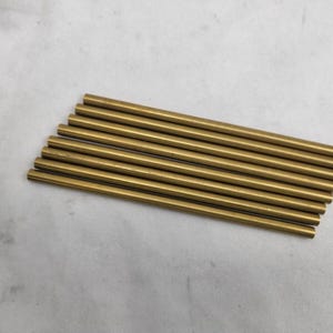 May include: A collection of ten gold-colored metal rods, each approximately 4 inches long, arranged diagonally on a light gray surface. The rods are cylindrical and appear to be made of brass or a similar alloy.