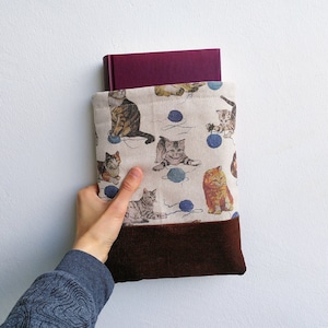Può includere: Una custodia per libri con tessuto beige a fantasia di gatti e gomitoli di lana blu. La parte inferiore è in velluto a coste marrone. Un libro bordeaux è all'interno.