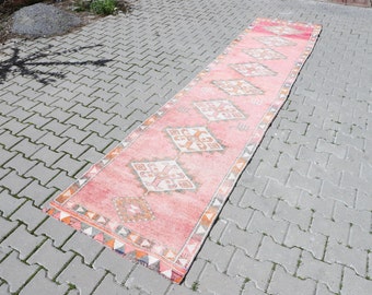 Puede incluir: Una alfombra de pasillo larga, rosa y naranja con un patrón geométrico. La alfombra tiene un aspecto descolorido y vintage y está colocada sobre un patio de adoquines grises.