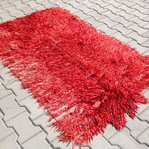 May include: A red shaggy rug with a long fringe on all sides. The rug is laid out on a gray concrete patio.