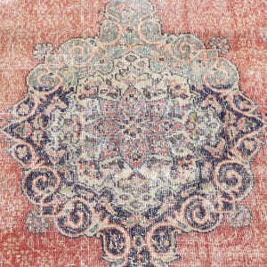 Pode incluir: Um tapete retangular com um fundo laranja terracota desbotado e um grande medalh&atilde;o central em azul-marinho, creme e verde-s&aacute;lvia. O padr&atilde;o intrincado inclui motivos florais sim&eacute;tricos e detalhes de vinhas entrela&ccedil;adas.