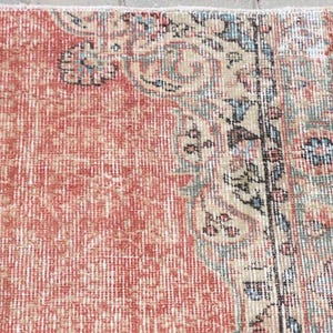 Pode incluir: Um tapete tecido com um grande campo central de vermelho terracota desbotado com uma textura desgastada. A borda exibe motivos florais e de vinhas intrincados em tons suaves de verde s&aacute;lvia, creme, azul claro e detalhes em cinza escuro.