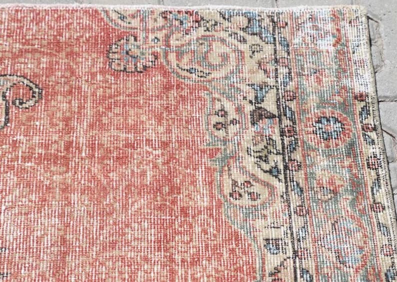Pode incluir: Um tapete tecido com um grande campo central de vermelho terracota desbotado com uma textura desgastada. A borda exibe motivos florais e de vinhas intrincados em tons suaves de verde s&aacute;lvia, creme, azul claro e detalhes em cinza escuro.