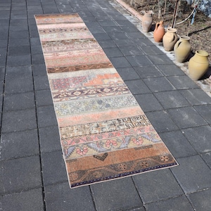 Peut inclure: Un long tapis de couloir rectangulaire présentant un motif patchwork composé de diverses bandes horizontales. Le tapis affiche une palette de couleurs douces comprenant du marron, orange, rose, gris et beige, avec des motifs géométriques et floraux.