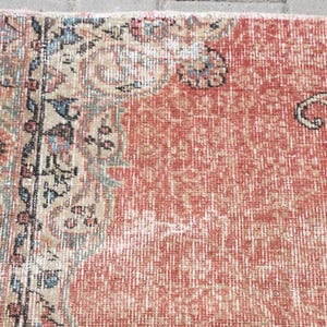 Pode incluir: Um tapete retangular tecido com um campo vermelho terracota proeminente e textura desgastada. A borda exibe padr&otilde;es florais e geom&eacute;tricos intrincados em tons suaves de verde s&aacute;lvia, creme, azul marinho e cinza carv&atilde;o.
