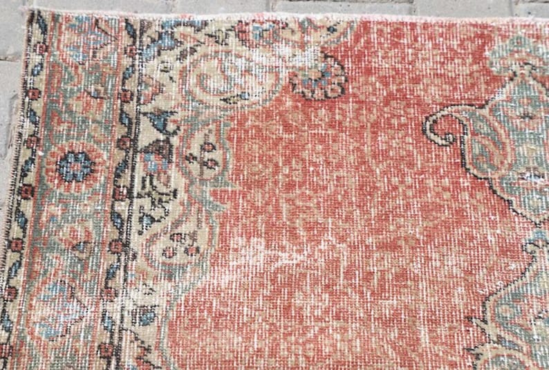 Pode incluir: Um tapete retangular tecido com um campo vermelho terracota proeminente e textura desgastada. A borda exibe padr&otilde;es florais e geom&eacute;tricos intrincados em tons suaves de verde s&aacute;lvia, creme, azul marinho e cinza carv&atilde;o.
