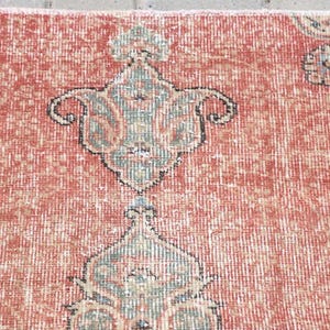 Pode incluir: Um tapete tecido com um fundo vermelho terracota proeminente e um padr&atilde;o repetitivo de motivos florais estilizados em tons de verde s&aacute;lvia, creme e azul escuro. O t&ecirc;xtil apresenta um pelo denso e texturizado com n&oacute;s e fibras vis&iacute;veis.