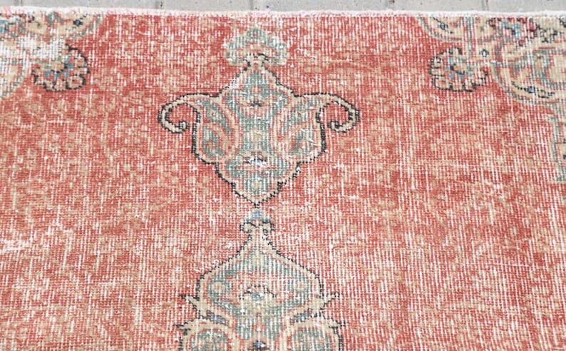 Pode incluir: Um tapete tecido com um fundo vermelho terracota proeminente e um padr&atilde;o repetitivo de motivos florais estilizados em tons de verde s&aacute;lvia, creme e azul escuro. O t&ecirc;xtil apresenta um pelo denso e texturizado com n&oacute;s e fibras vis&iacute;veis.