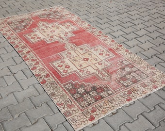 Puede incluir: Una alfombra rectangular roja desgastada con un diseño geométrico beige central y un borde estampado. La alfombra tiene una apariencia descolorida, lo que sugiere que es vintage o tiene una estética desgastada. La alfombra está colocada sobre una superficie de ladrillos grises.
