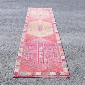 May include: A rectangular, vintage-style rug with a geometric design. The rug features a color palette of pink, peach, and cream, with a central pattern of diamonds and rectangles. The border has a checkered pattern.