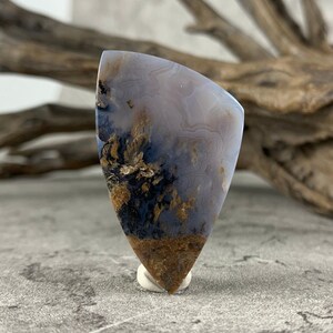 May include: A teardrop-shaped agate cabochon with a white, gray, and brown color pattern. The agate has a unique, swirling pattern with brown dendrites.