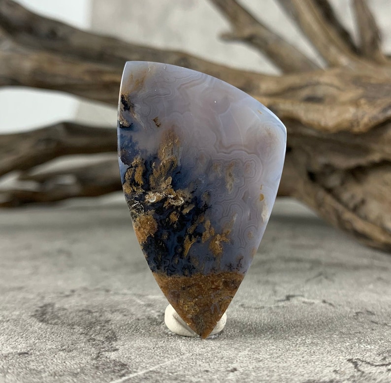 May include: A teardrop-shaped agate cabochon with a white, gray, and brown color pattern. The agate has a unique, swirling pattern with brown dendrites.