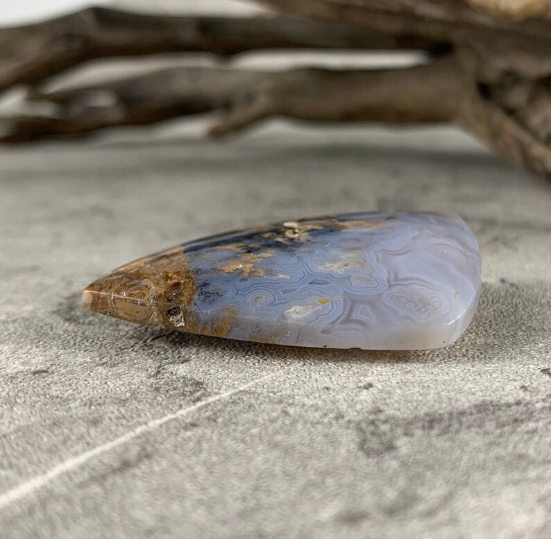 May include: A light blue and brown agate cabochon with a swirling pattern. The cabochon is shaped like a teardrop and has a smooth, polished surface.