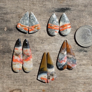 Op de afbeelding: Een assortiment gepolijste edelsteen cabochons in diverse vormen, waaronder druppels en driehoeken. De stenen hebben een mix van grijze, oranje, witte en bruine kleuren. Een Amerikaanse kwart dollar munt is inbegrepen ter referentie.