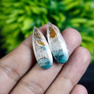 Puede incluir: Dos piedras preciosas en forma de lágrima con un acabado pulido. Las piedras presentan una mezcla de colores, incluyendo turquesa, blanco, marrón y gris. Las piedras preciosas se sostienen en una mano, con un fondo verde borroso.