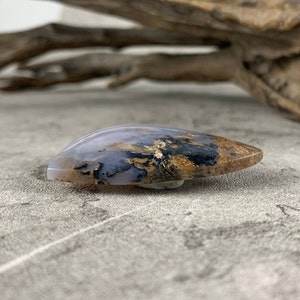 May include: A teardrop-shaped agate cabochon with a white, gray, and brown pattern. The agate has a smooth, polished surface.