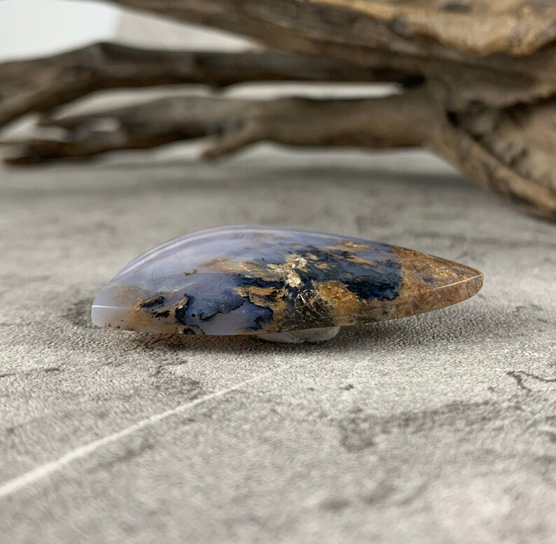 May include: A teardrop-shaped agate cabochon with a white, gray, and brown pattern. The agate has a smooth, polished surface.