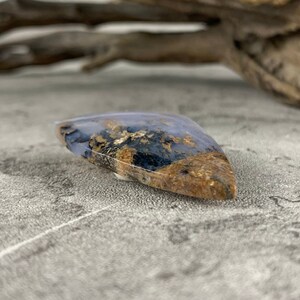 May include: A teardrop-shaped cabochon of blue agate with gold and black inclusions. The stone is polished and has a smooth, glossy finish.