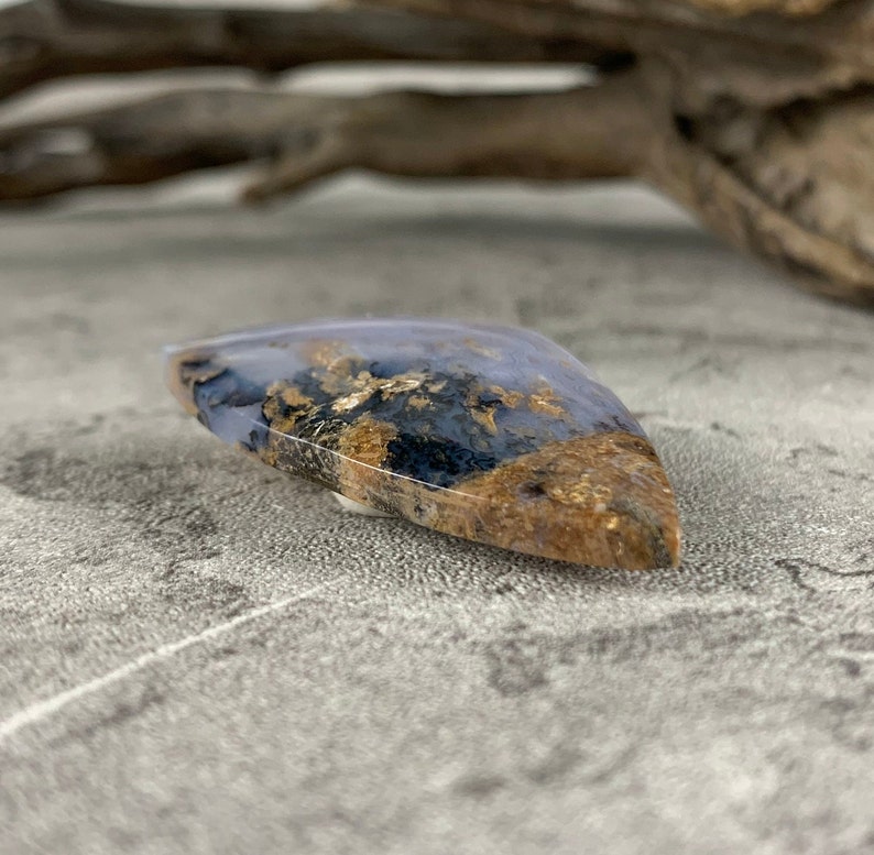 May include: A teardrop-shaped cabochon of blue agate with gold and black inclusions. The stone is polished and has a smooth, glossy finish.