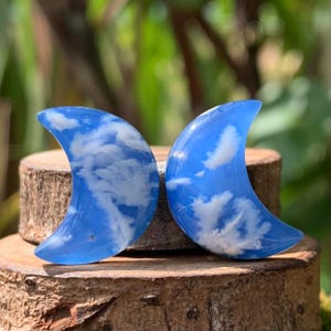 May include: A pair of crescent moon shaped earrings with a blue resin base and white clouds embedded inside.