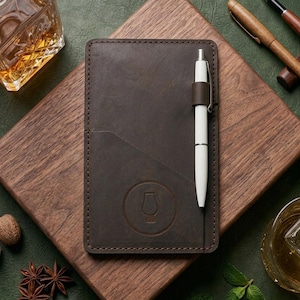 May include: A brown leather notebook with a white pen, a glass of whisky with ice, and two wooden pens on a wooden board. The scene includes a bottle of liquor, spices, and mint leaves, all arranged on a green surface.