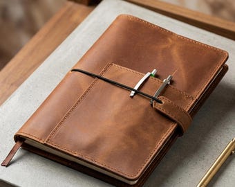 May include: A brown leather journal with a pen holder and a black elastic band closure. Two pens are secured in the holder. A gold pen rests on the surface next to the journal. The journal is on a gray surface.