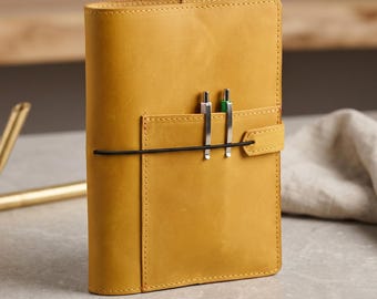 May include: A mustard-yellow leather journal with a pen holder and a black elastic closure. Two pens, one silver and one green, are in the pen holder. A gold pen holder and a white cloth are in the background.