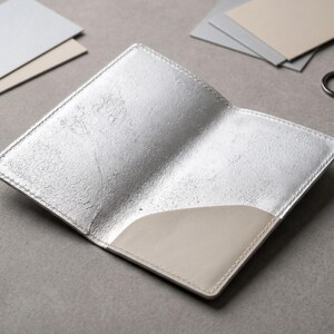 May include: Open silver leather passport holder with a light beige pocket. The passport holder is open, revealing the interior. Two small silver key rings and a few small cards are visible in the background. The passport holder is on a gray surface.