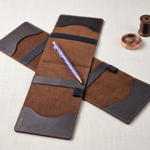 May include: Open brown leather pen case with a blue and white patterned pen inside. The case has dark brown and black accents, with a dark blue strap. A spool of thread and a compass are in the background.