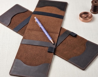 May include: Open brown leather pen case with a blue and white patterned pen inside. The case has dark brown and black accents, with a dark blue strap. A spool of thread and a compass are in the background.