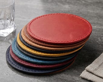 May include: A collection of oval leather coasters in various colours, including red, brown, and blue. Each coaster has a stitched edge. A clear glass and a folded linen napkin are also visible.