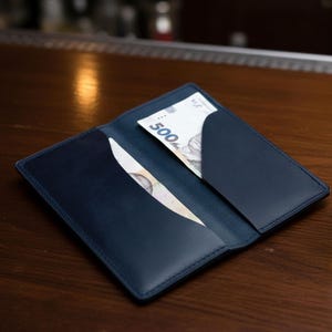 May include: A navy blue leather wallet lies open on a wooden surface, with banknotes visible in its pockets. A shot glass containing amber liquid is positioned to the right of the wallet.