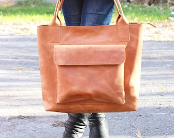 May include: A brown leather tote bag with a large front pocket. The bag has a zipper closure and two top handles.