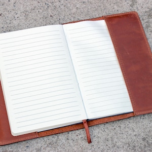 K&ouml;nnte beinhalten: Offenes Notizbuch mit linierten Seiten, umh&uuml;llt von einem braunen Ledercover. Das Notizbuch ist ge&ouml;ffnet und zeigt leere, linierte Seiten. Ein braunes Lesezeichenband ist vorhanden. Das Notizbuch liegt auf einer grauen Oberfl&auml;che.