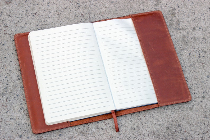 K&ouml;nnte beinhalten: Offenes Notizbuch mit linierten Seiten, umh&uuml;llt von einem braunen Ledercover. Das Notizbuch ist ge&ouml;ffnet und zeigt leere, linierte Seiten. Ein braunes Lesezeichenband ist vorhanden. Das Notizbuch liegt auf einer grauen Oberfl&auml;che.